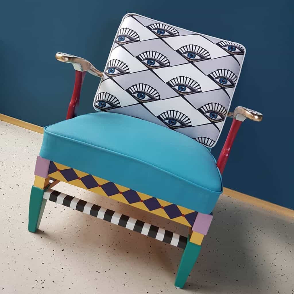 Whimsical armchair with a bold eye-patterned backrest, turquoise seat, and multicolored hand-painted legs for a playful artistic touch