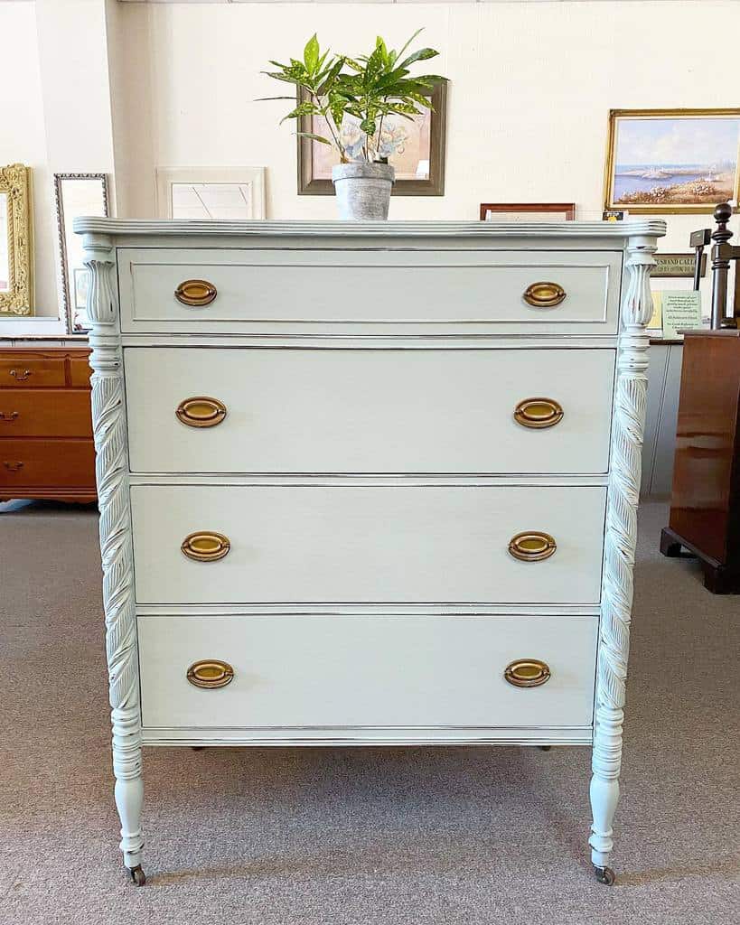 Classic vintage dresser refinished in soft white chalk paint with carved details, brass oval handles, and elegant spindle legs on casters