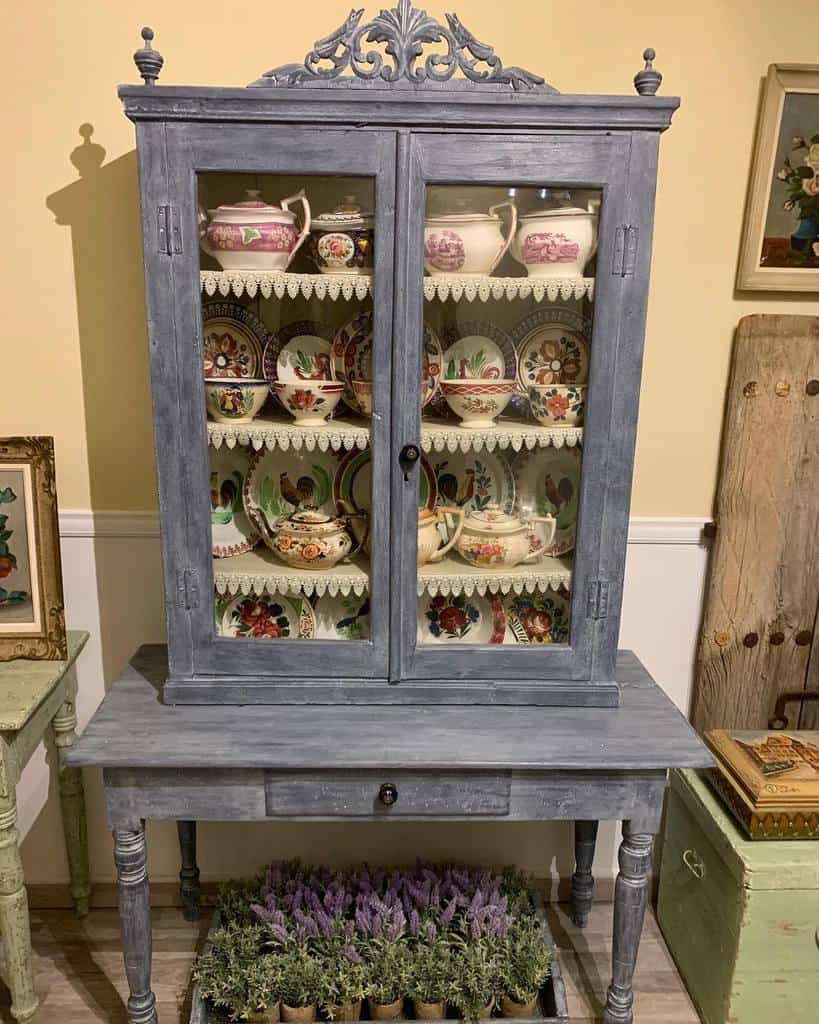 Antique hutch with distressed blue chalk paint, ornate detailing, and glass doors displaying vintage china for a charming rustic look