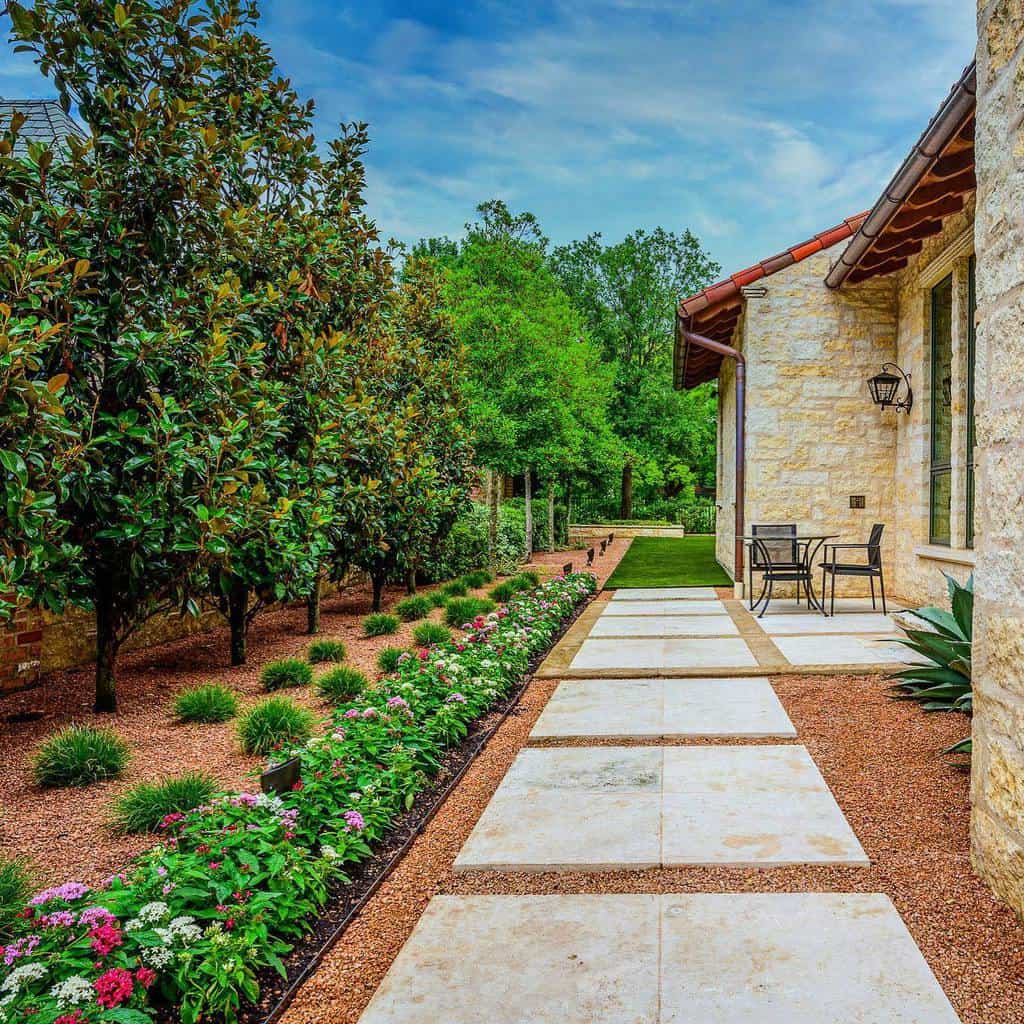 Elegant xeriscape walkway with large stone pavers, lush greenery, flowering plants, and neatly trimmed trees alongside a stone house