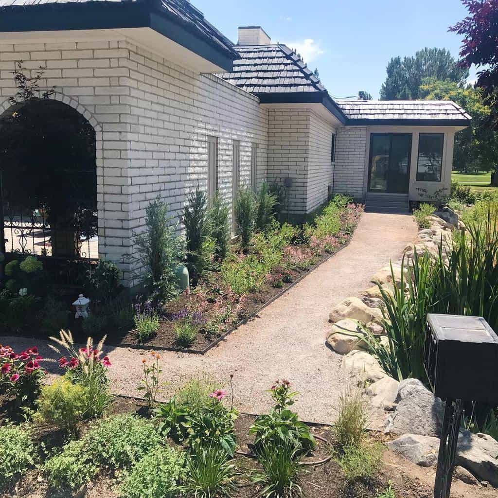 A scenic xeriscape walkway with drought-tolerant plants, decorative rocks, and a curved gravel path leading to a white brick house