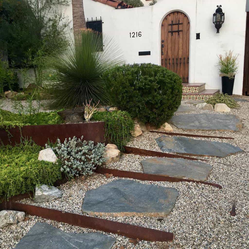 A Mediterranean-style xeriscape entrance with a rustic wooden door, gravel ground cover, large stone pavers, drought-tolerant plants, and corten steel edging