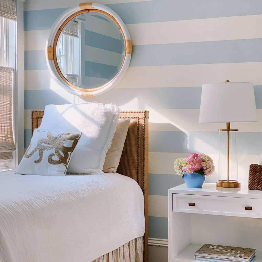 Coastal-themed bedroom with blue and white striped walls, round mirror, white bed, octopus pillow, and a lamp on a white nightstand with flowers