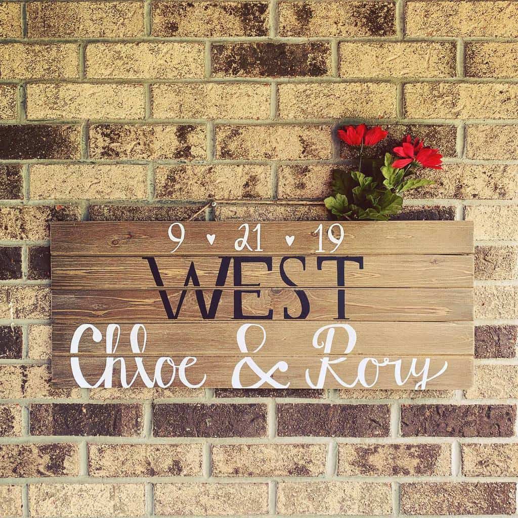 Wooden sign on brick wall with "West," "Chloe & Roy," and date "9-21-19" Red flowers are attached at the top