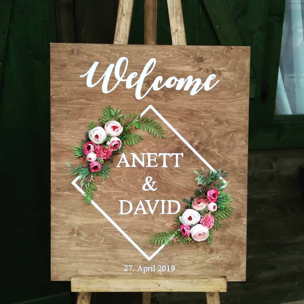 Wooden wedding sign with "Welcome Anett & David" in white, decorated with pink and white flowers. Date: 27 April 2019