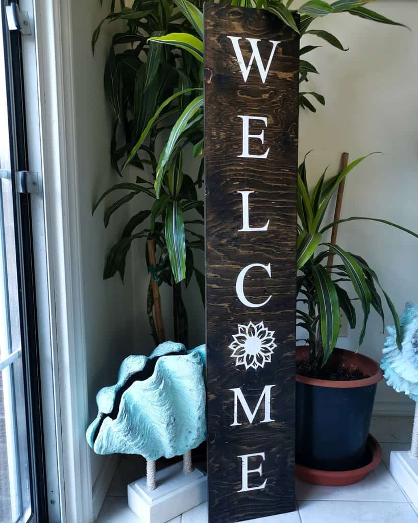 A wooden sign with "WELCOME" in white letters stands next to potted plants and decorative teal shells indoors