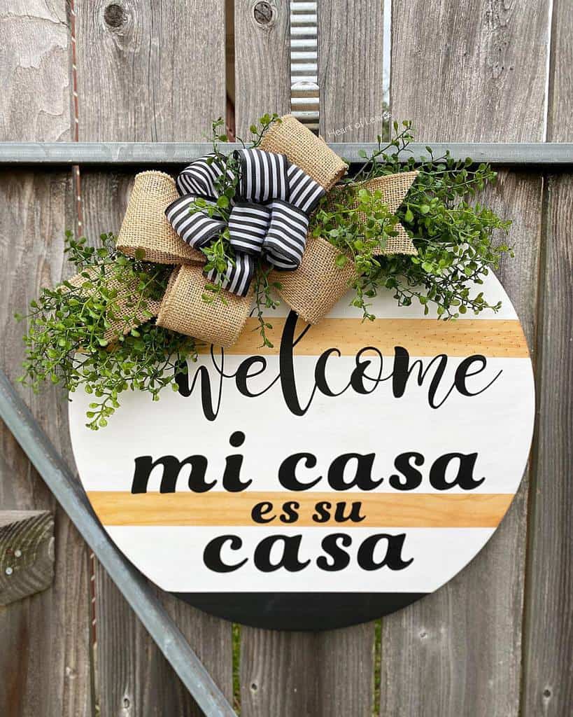 Round wooden sign on a fence reads "Welcome, mi casa es su casa," decorated with burlap and striped ribbons and greenery