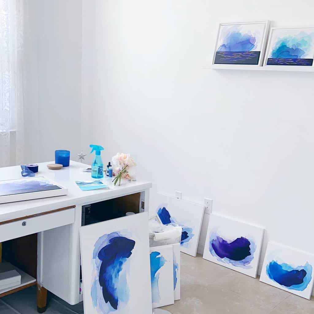 Art studio with abstract blue and purple paintings on canvases, a desk with art supplies, and paintings displayed on a shelf