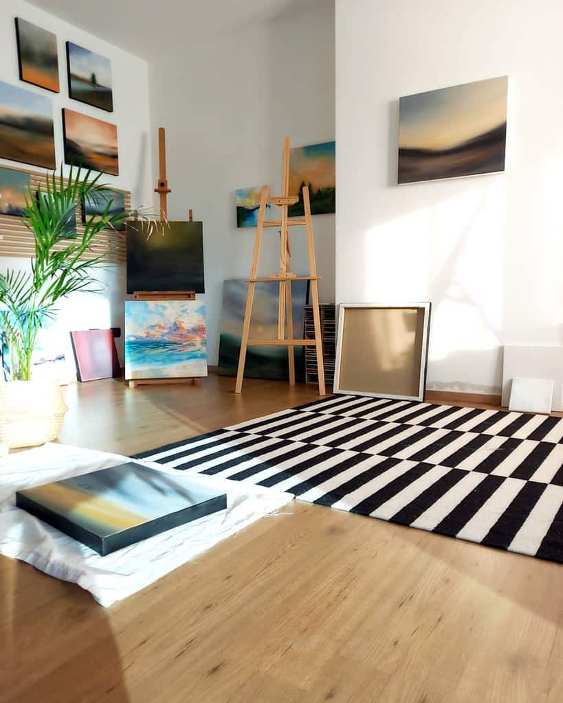 Art studio with nature paintings on walls and easels, a striped rug, and a potted plant in a sunlit room with wooden floor