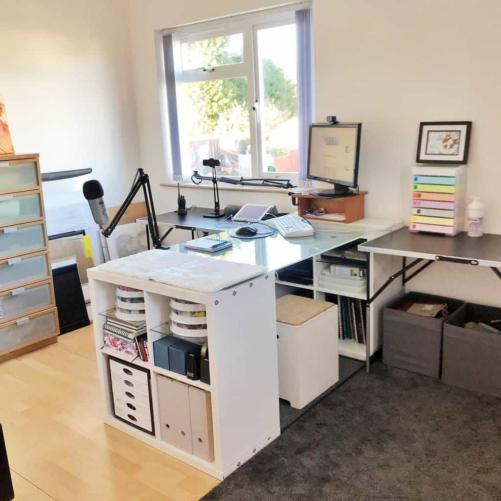 Bright and organized art studio with a white desk, storage shelves, a microphone, and a computer setup near a large window