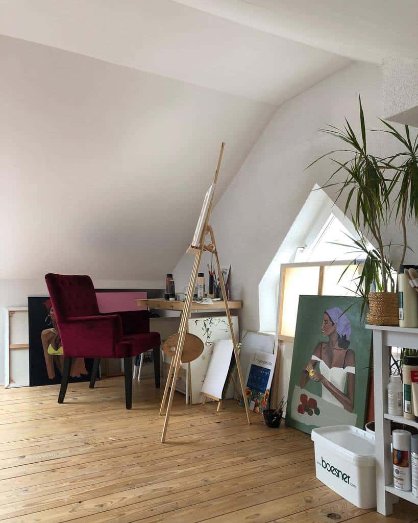 Bright attic art studio with an easel, paintings, a red chair, and a potted plant by a skylight window