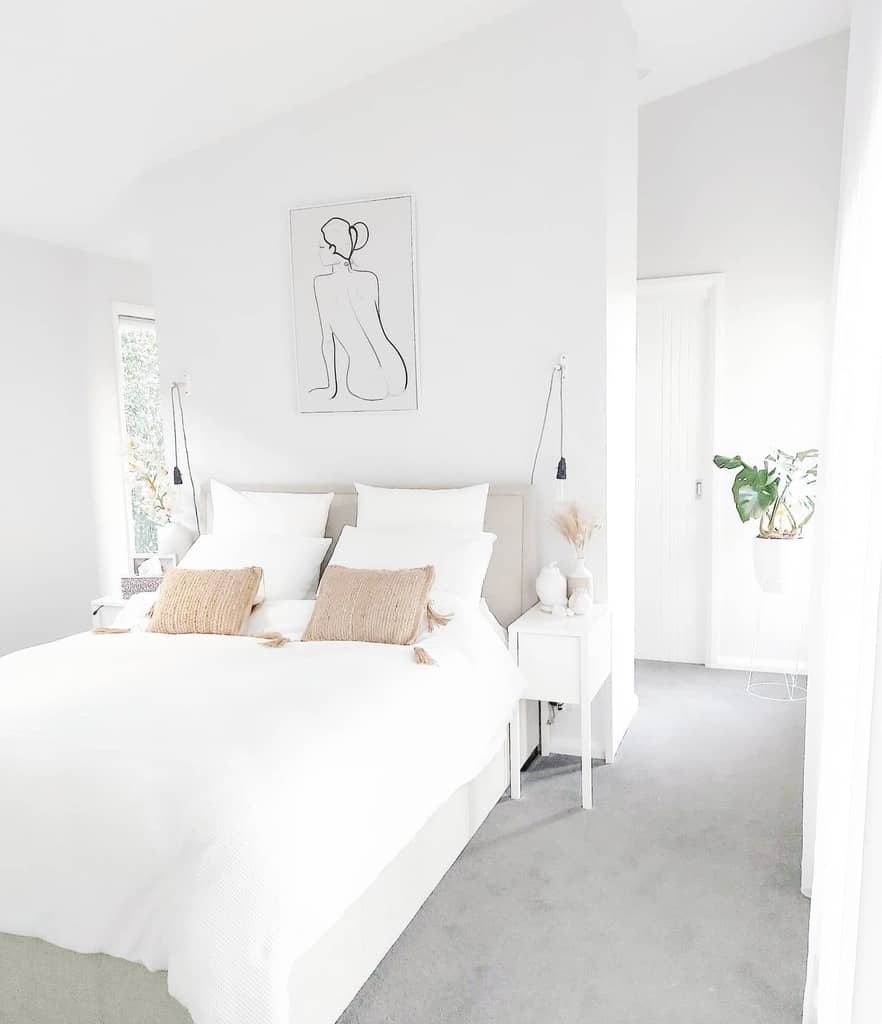 Minimalist white bedroom with a bed, beige pillows, abstract wall art, and a plant on a side table creating a serene atmosphere