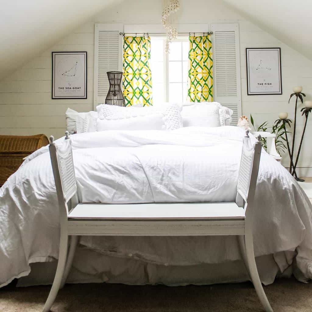 Cozy attic bedroom with white bedding, green patterned curtains, and zodiac-themed wall art; white bench at the foot of the bed