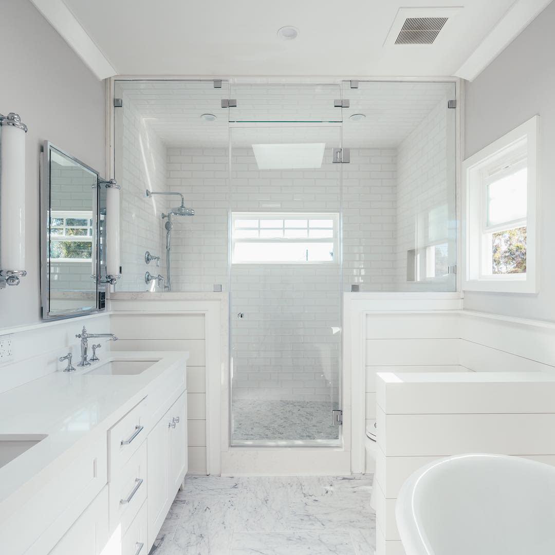 Elegant all-white wet room with a glass-enclosed shower, marble flooring, double vanity, and bright natural light for a fresh, airy feel