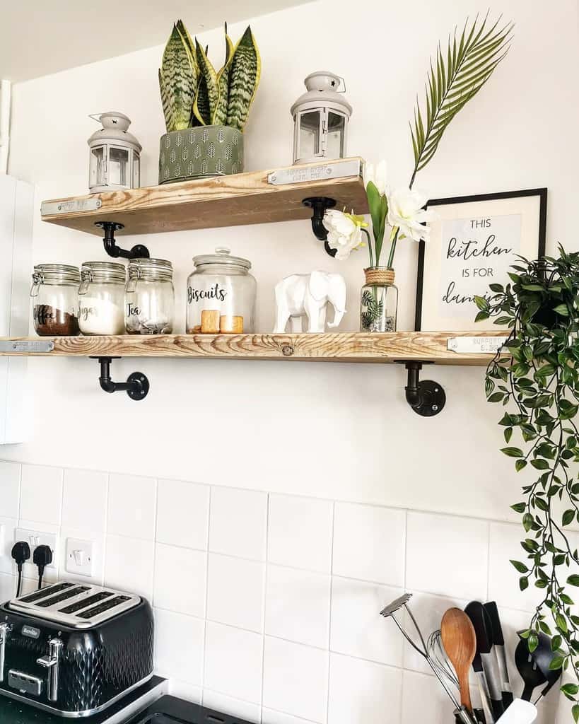 Kitchen shelves with plants, lanterns, jars, framed sign, and utensils on a counter with white tiles and a stovetop