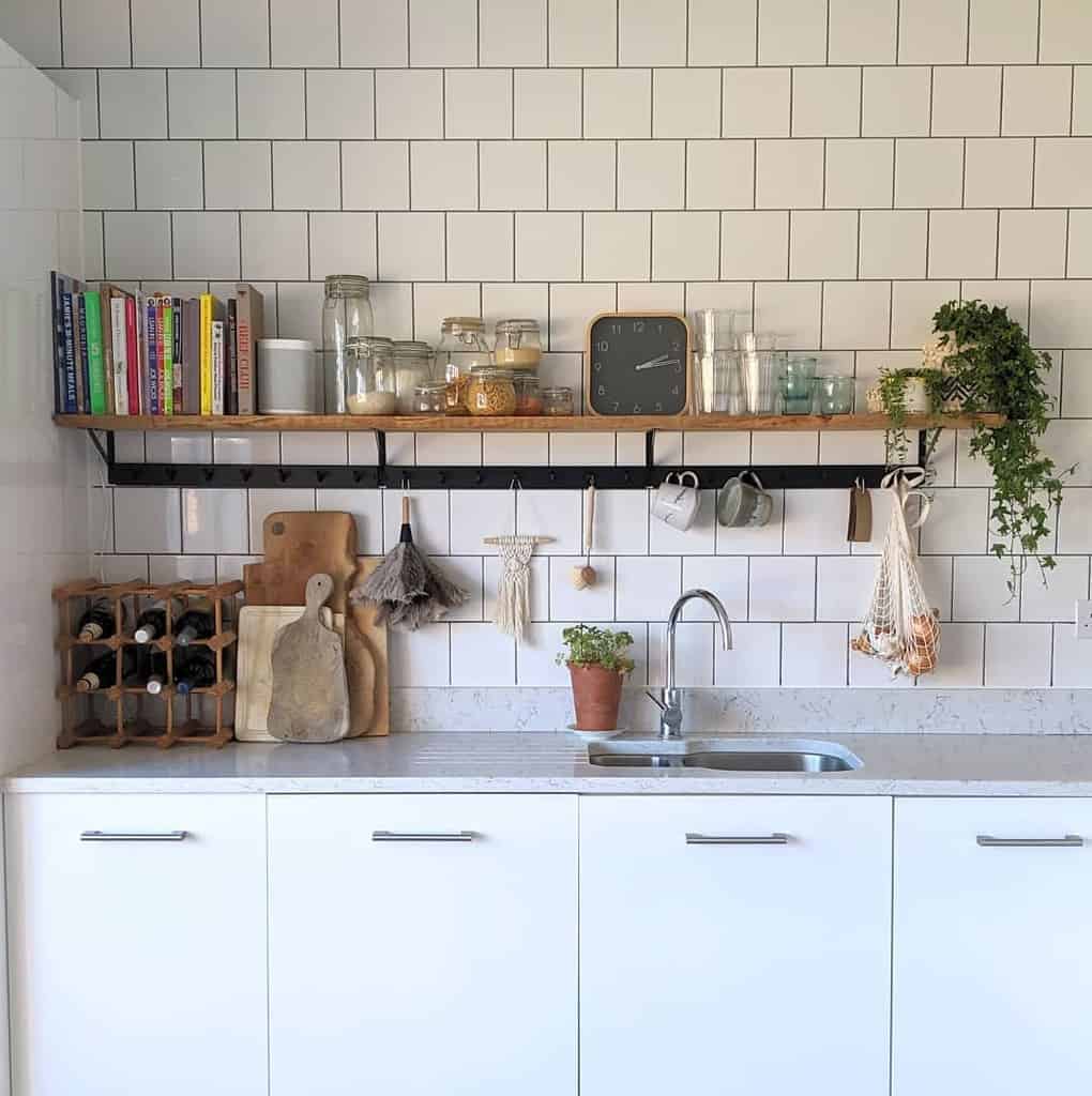 Wooden open shelf with black brackets holds books, jars, and plants, while hooks below store mugs and decor in this modern farmhouse kitchen