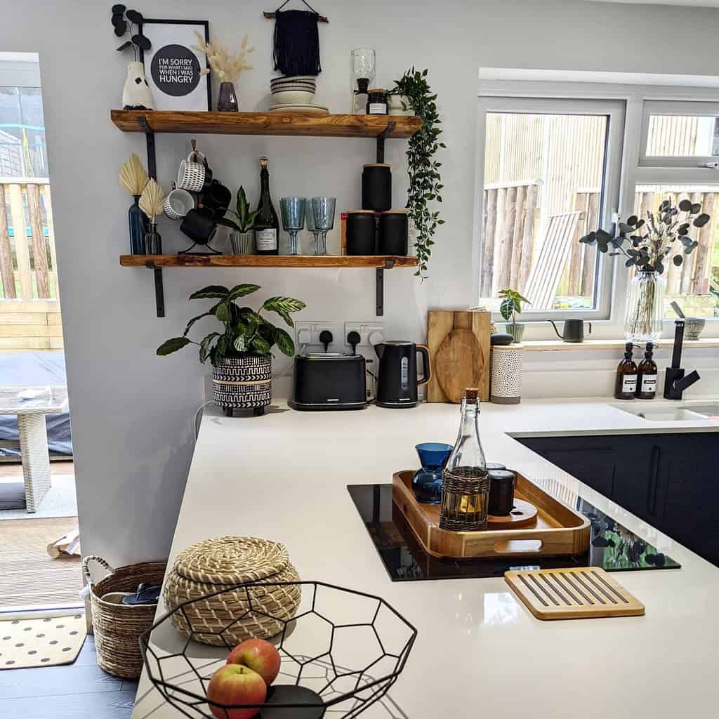 Rustic wooden shelves with black brackets display glassware, mugs, and plants, adding warmth and style to a modern kitchen with white countertops