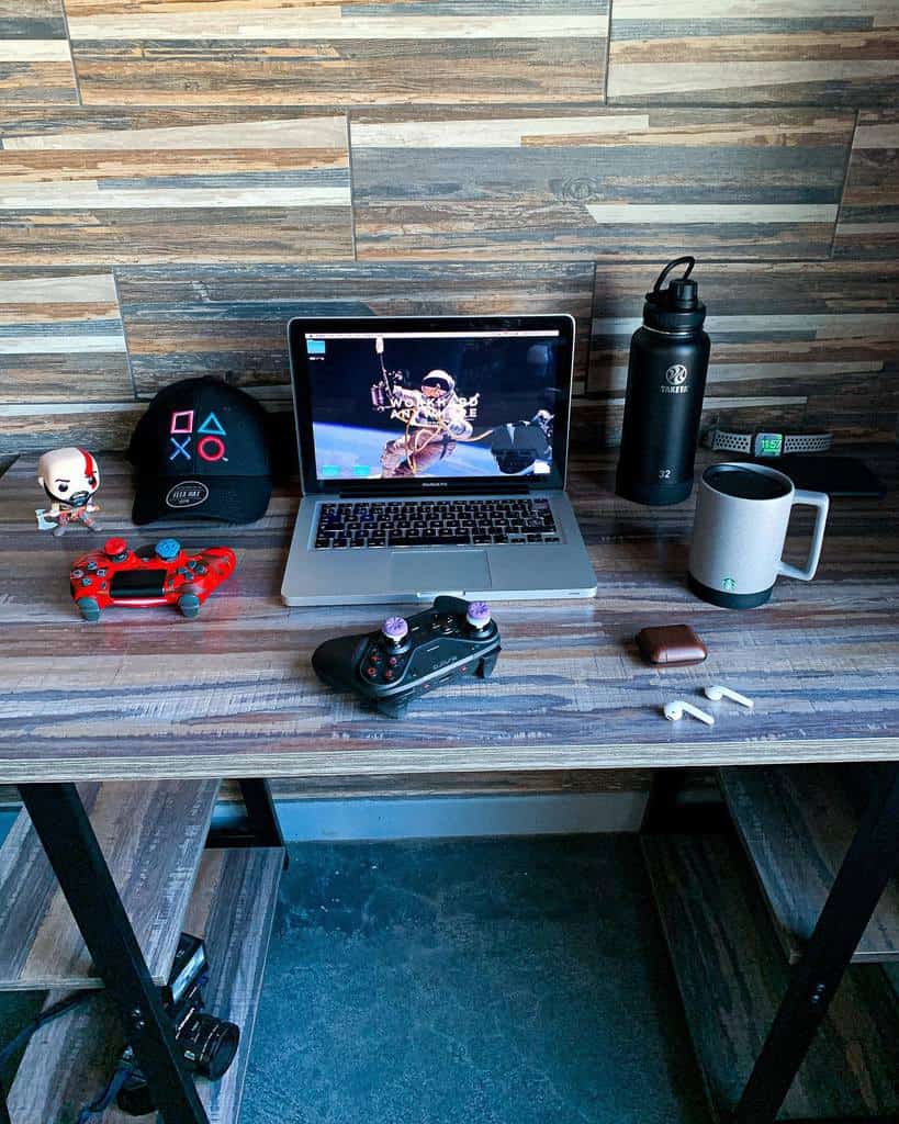 A gaming setup on a wood desk includes a laptop, red game controller, black cap, headphones, mugs, water bottle, and various accessories