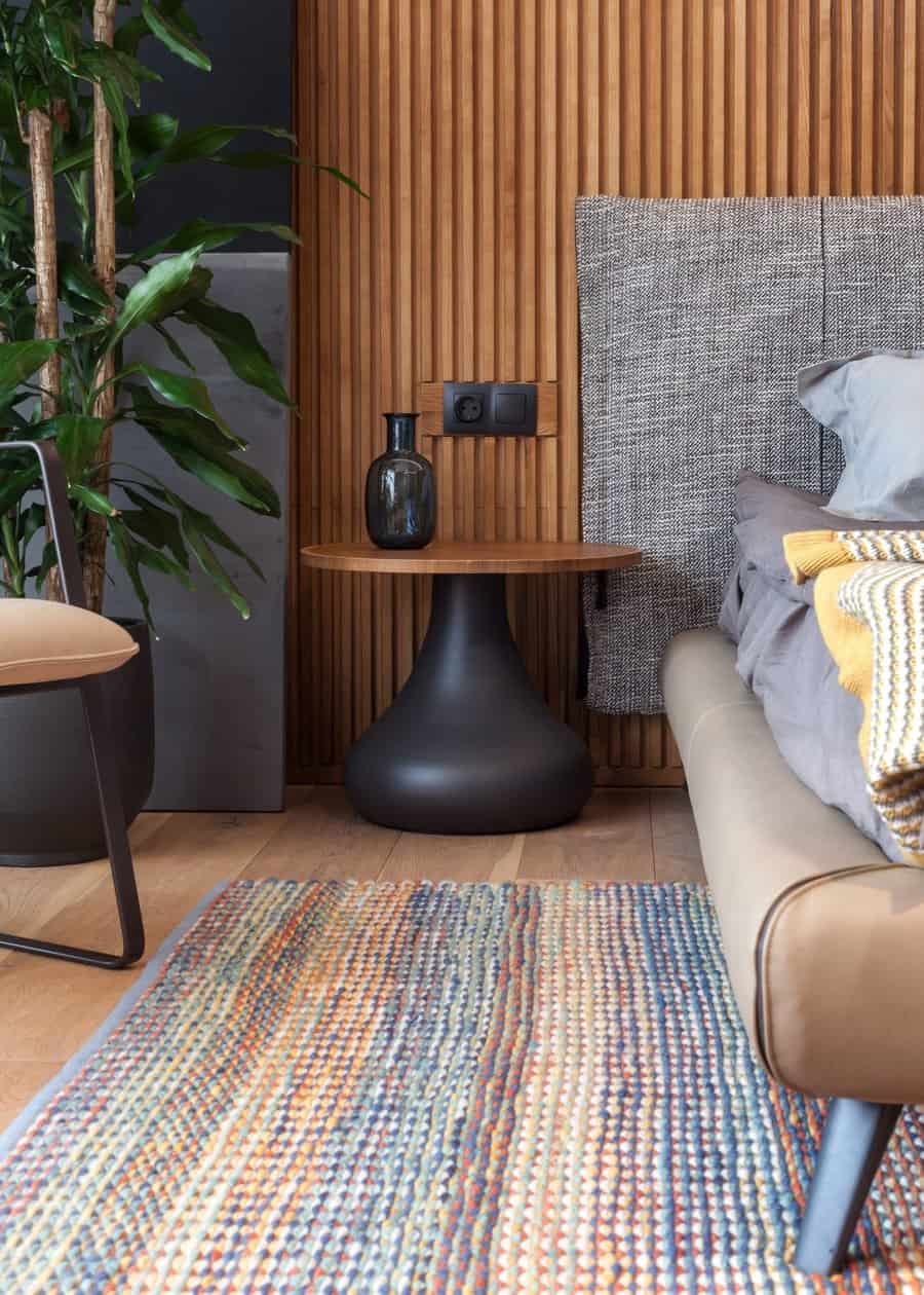 Cozy bedroom with a modern side table, plant, multicolored rug, and a gray bed; a wall with vertical wooden panels adds warmth