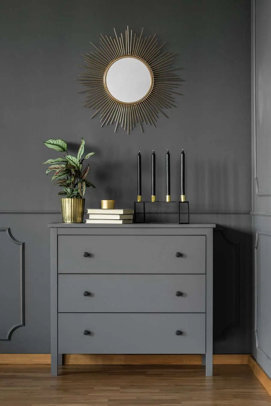 Gray dresser with decor items, gold mirror, plant, candles, and books on a gray wall background