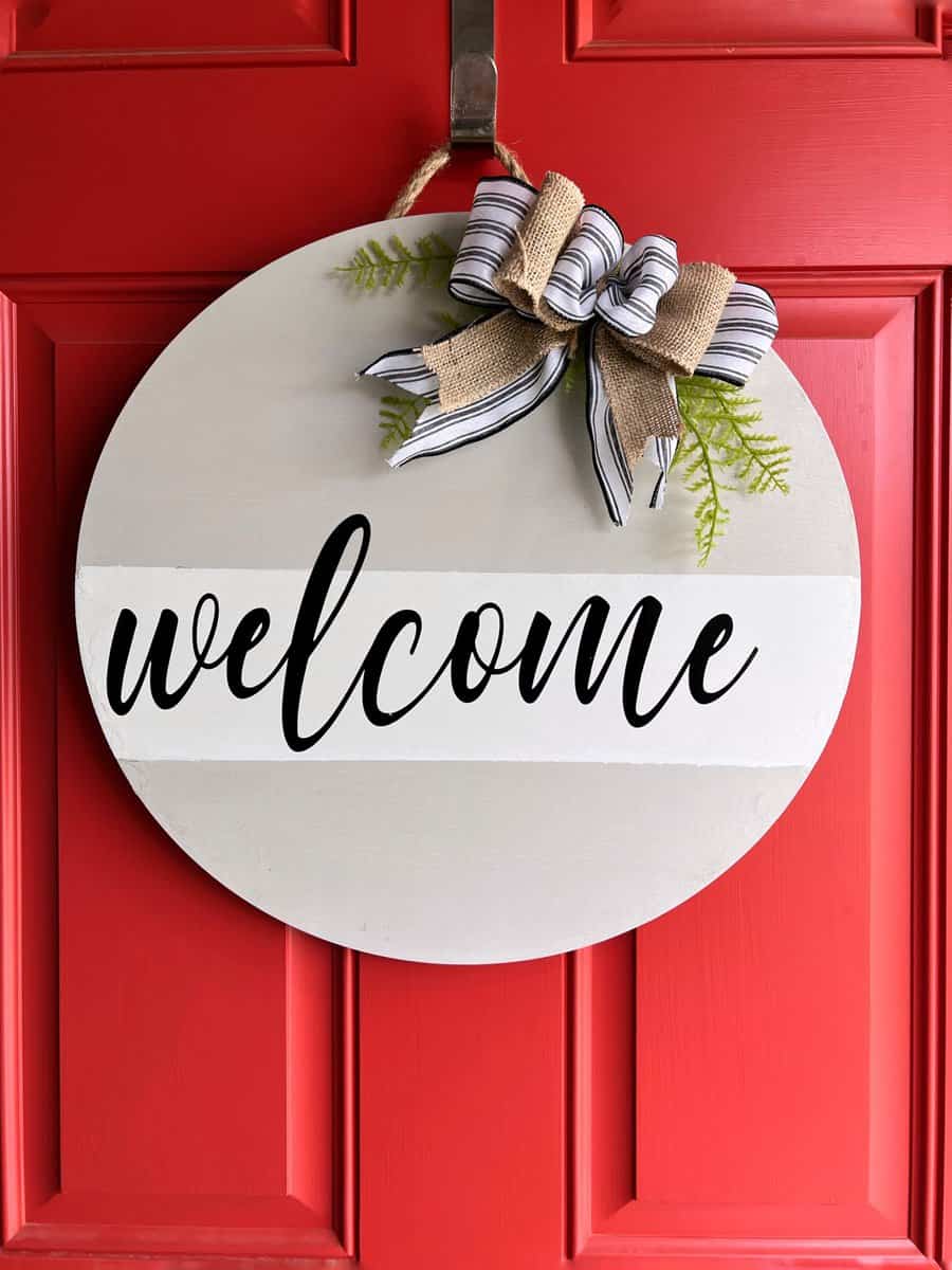 Round welcome sign with a striped bow and greenery hangs on a bright red door with paneled design