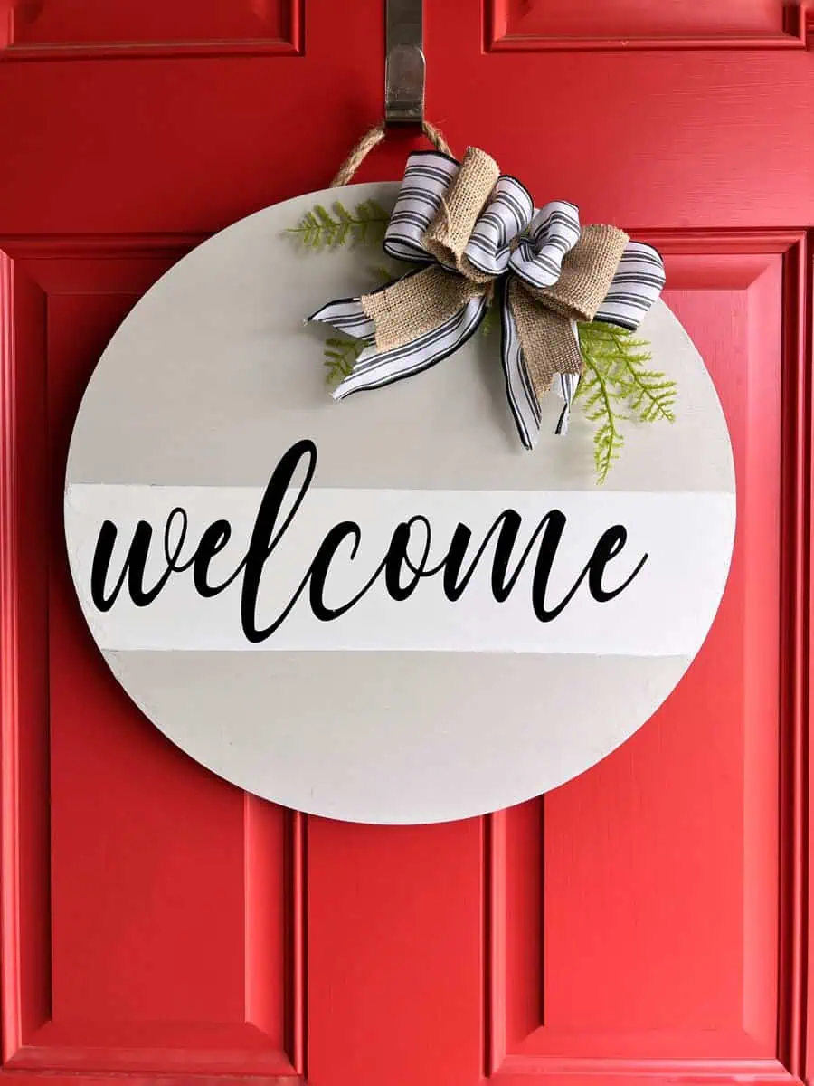 Round welcome sign with a striped bow and greenery hangs on a bright red door with paneled design.