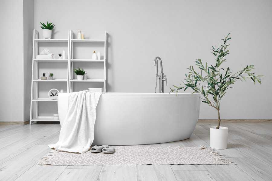 Minimalistic bathroom featuring a white oval bathtub, a plant, and shelves with a towel and slippers placed on a patterned rug on the light wood floor