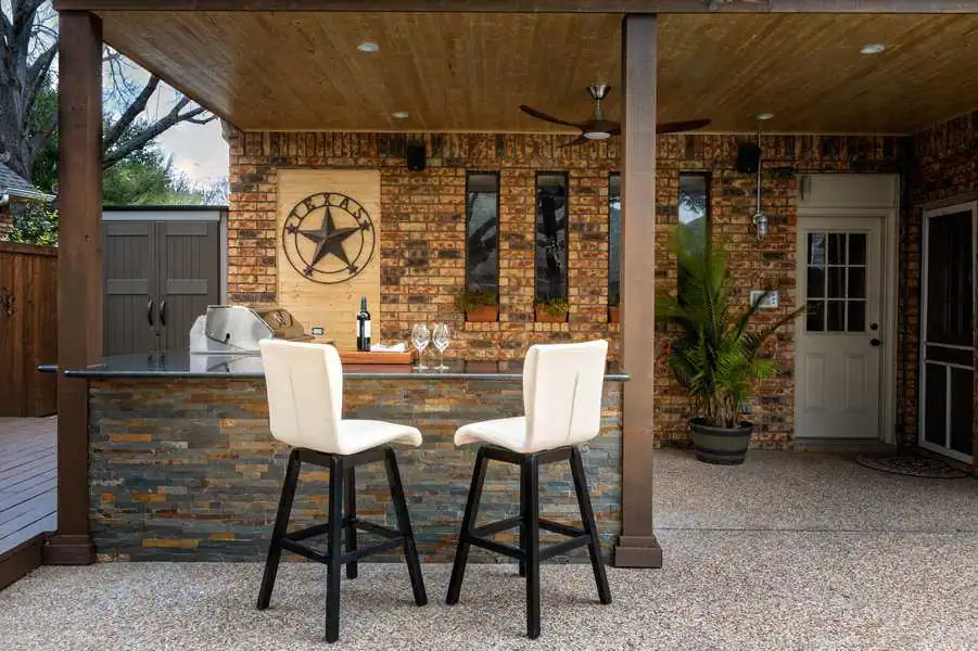 Outdoor kitchen featuring brick walls, two white bar stools, and a wooden pergola roof