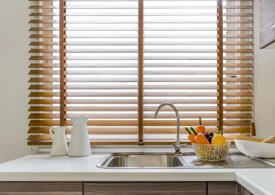 Modern kitchen sink with wooden blinds, a basket of fruits, and white jugs on the counter