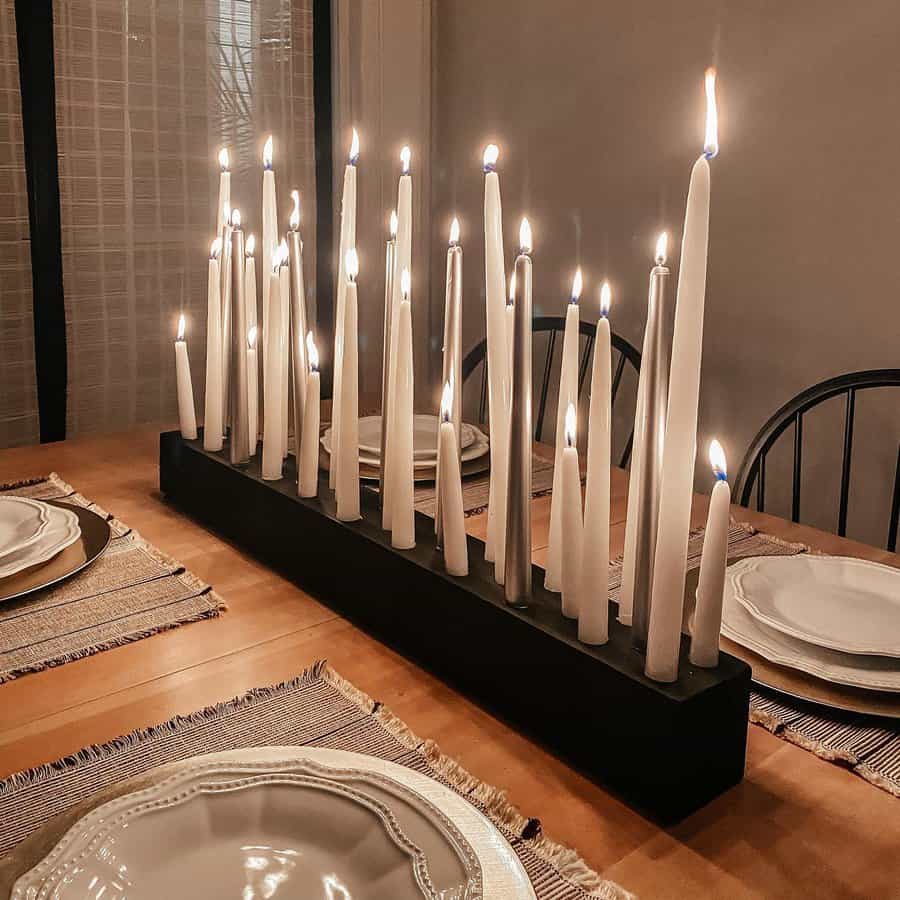 A wooden dining table set with four place settings and a centerpiece of numerous tall lit white candles arranged in a black holder