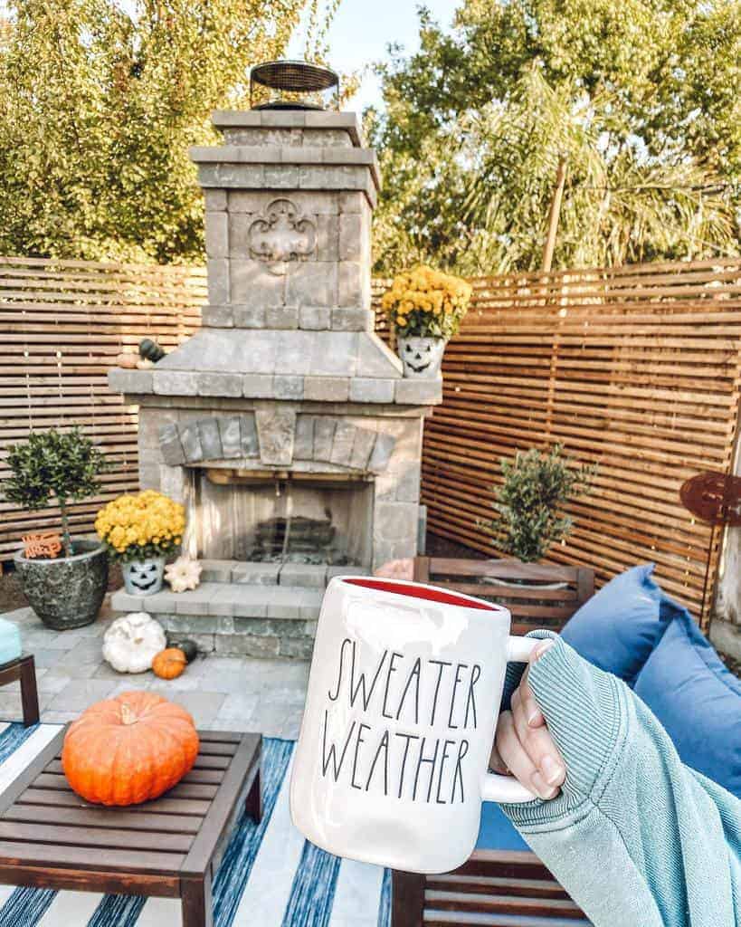 Cozy outdoor patio with a stone fireplace, wooden slat privacy screen, autumn decor, pumpkins, and a 'Sweater Weather' mug capturing the fall ambiance