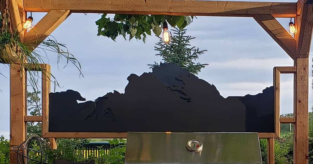 Outdoor grill area with a decorative privacy screen featuring a mountain silhouette, framed by a wooden pergola with hanging lights and lush greenery