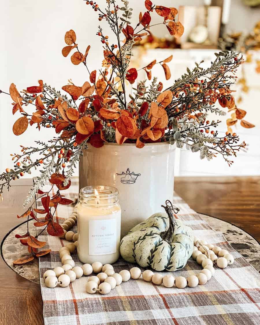 Ceramic pot with orange and red foliage, a lit candle, decorative pumpkin, and bead garland on a plaid tablecloth