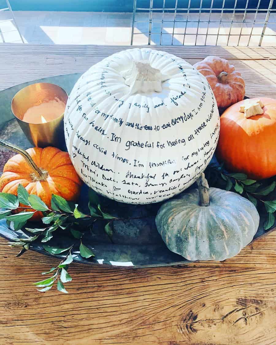 Decorative pumpkins on a tray, with one white pumpkin featuring handwritten messages and surrounded by candles and greenery