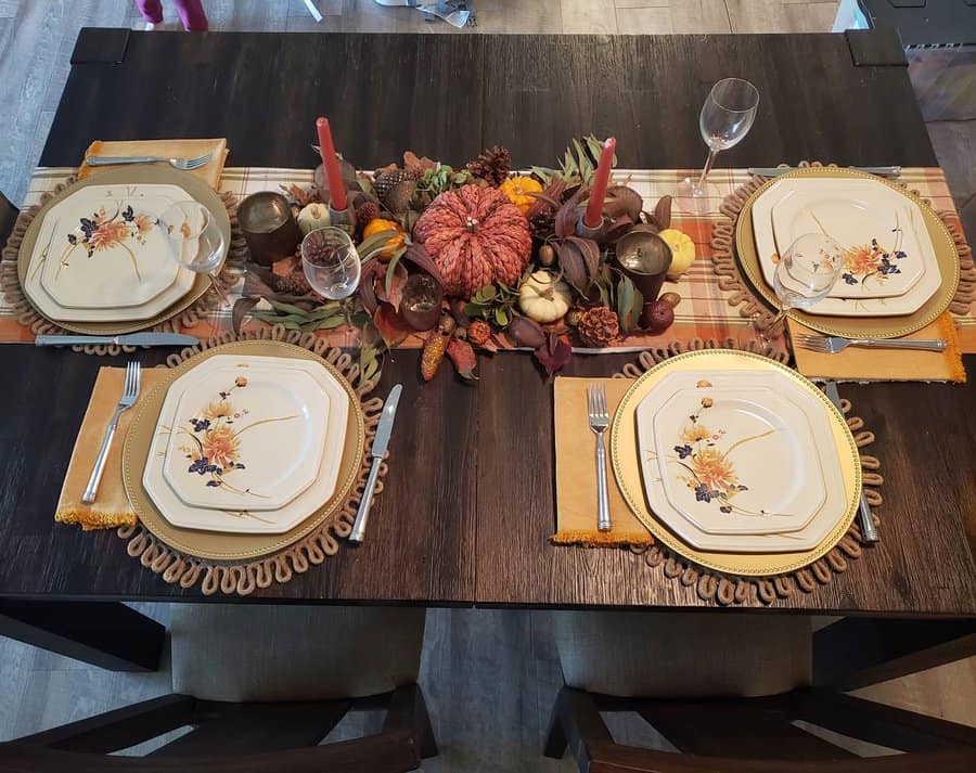 A fall-themed dining table is set for four with floral plates, orange candles, and a Thanksgiving centerpiece of gourds and leaves