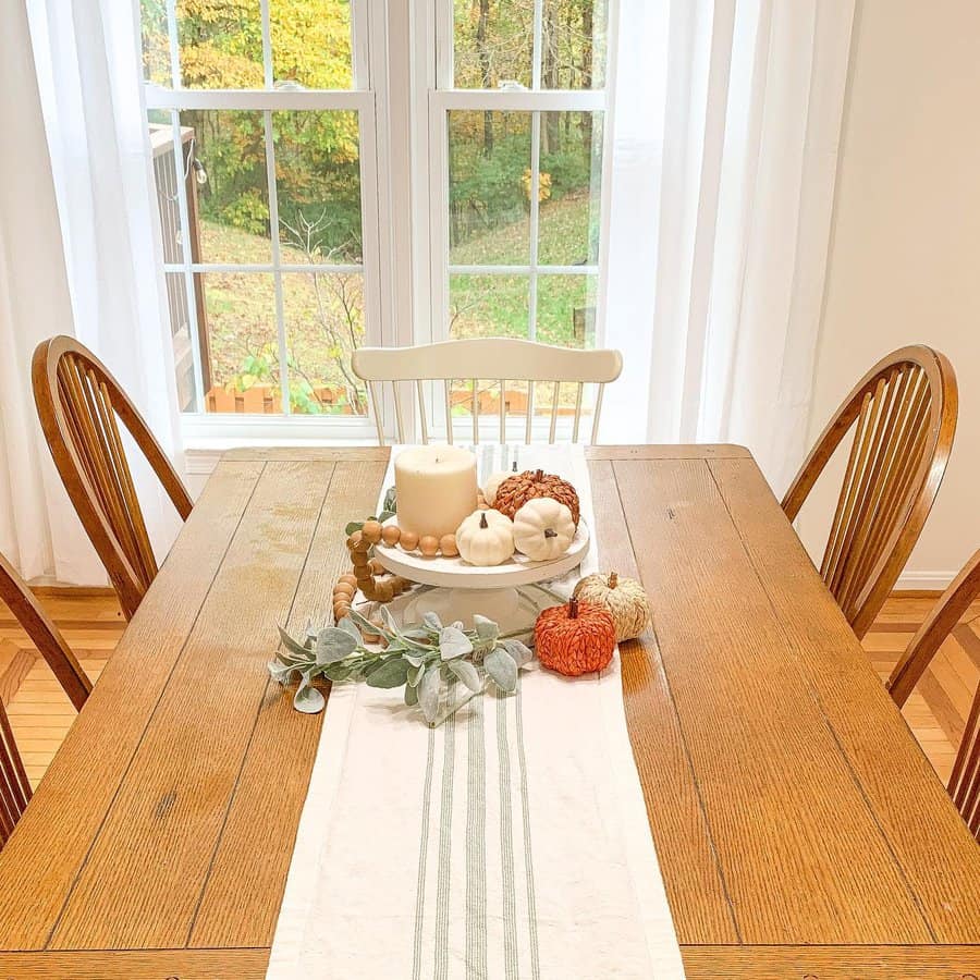 A dining table with fall decor, including small pumpkins, a white candle, and greenery, in front of large windows with sheer curtains