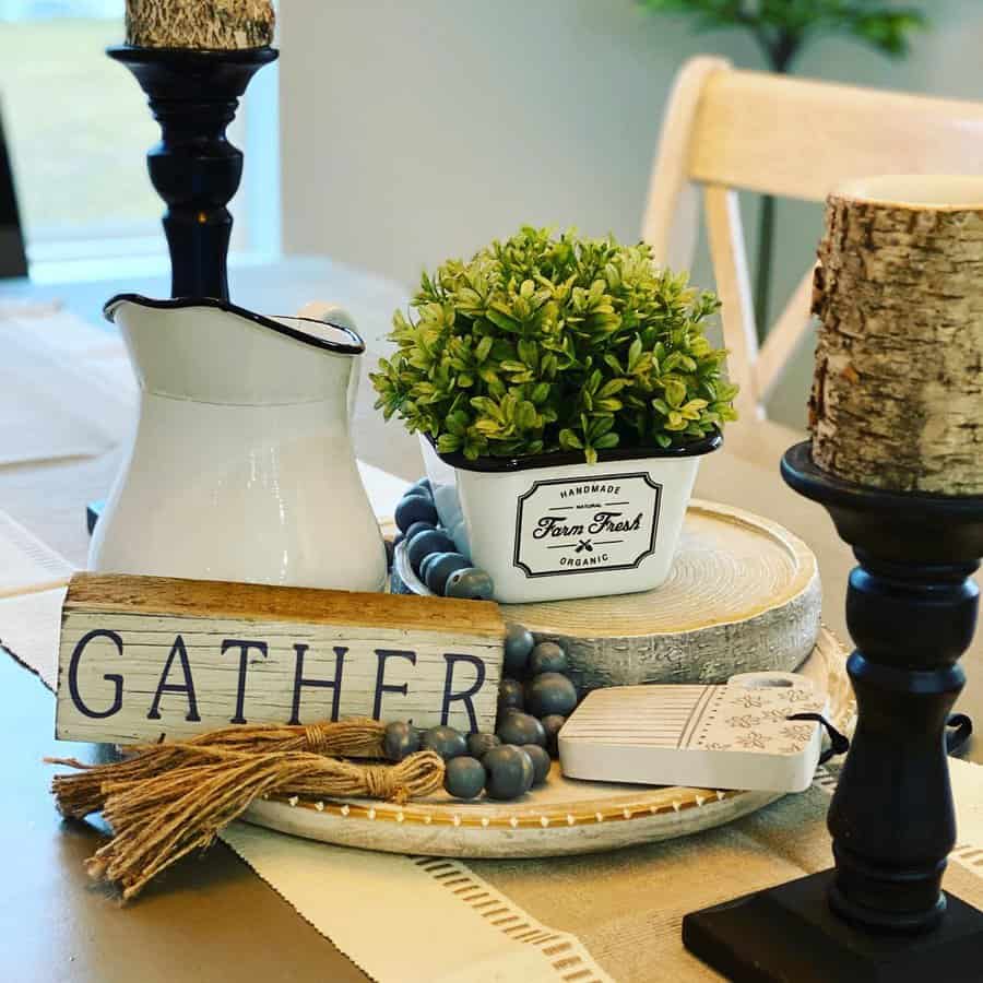 Rustic table centerpiece with a potted plant, a white pitcher, a "Gather" sign, wooden beads, and candles on a tray