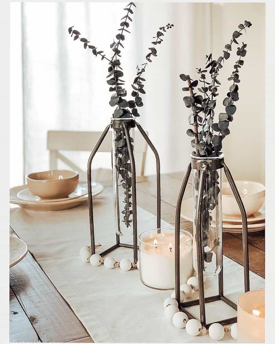 A dining table with eucalyptus in vases, candles, and ceramic dishes on a wooden surface with a beige runner, creating a cozy and minimalist atmosphere