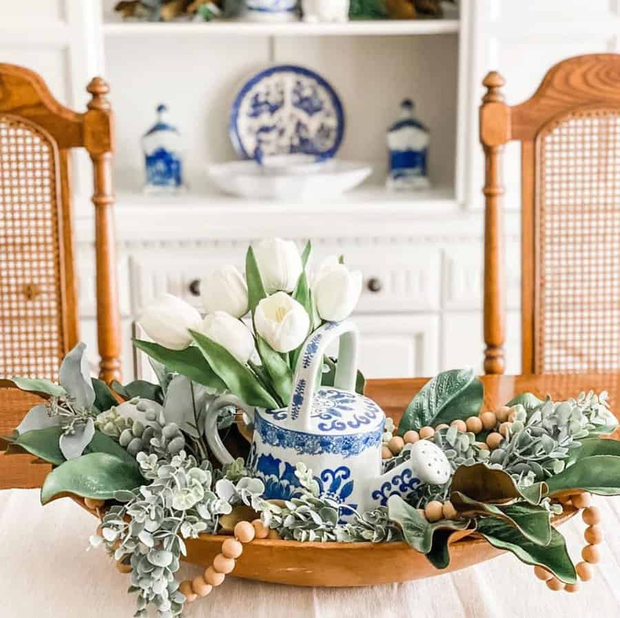 A wooden tray with white tulips in a blue and white teapot, surrounded by greenery and beads, on a dining table