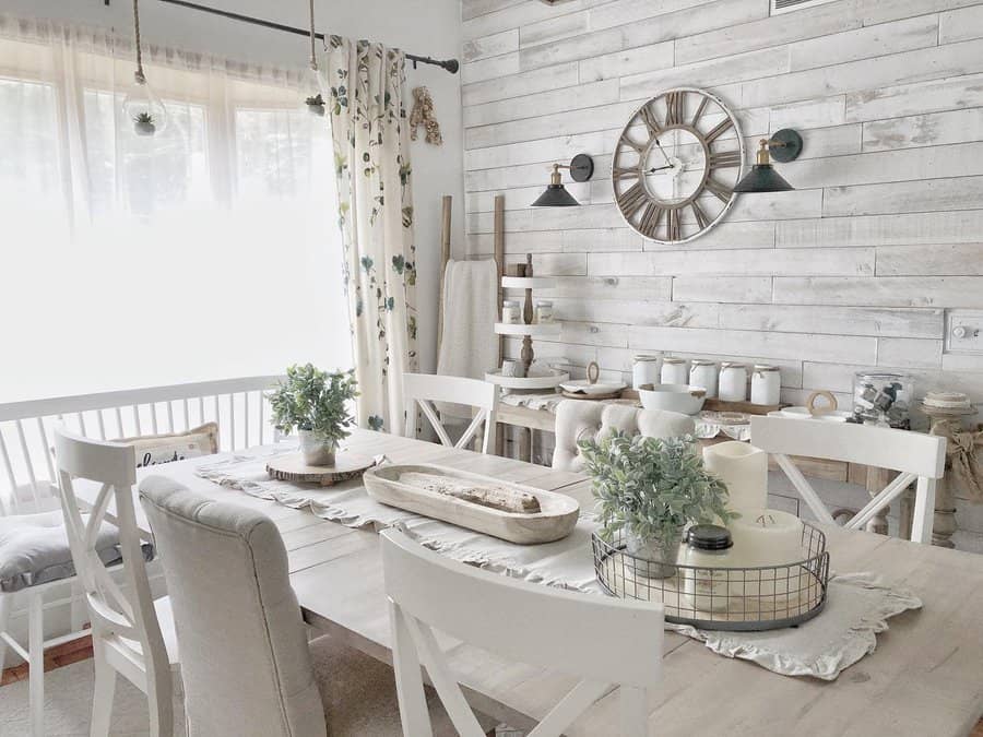 Bright farmhouse dining room with a rustic wood table, neutral decor, greenery, candles, and a vintage-inspired wall clock