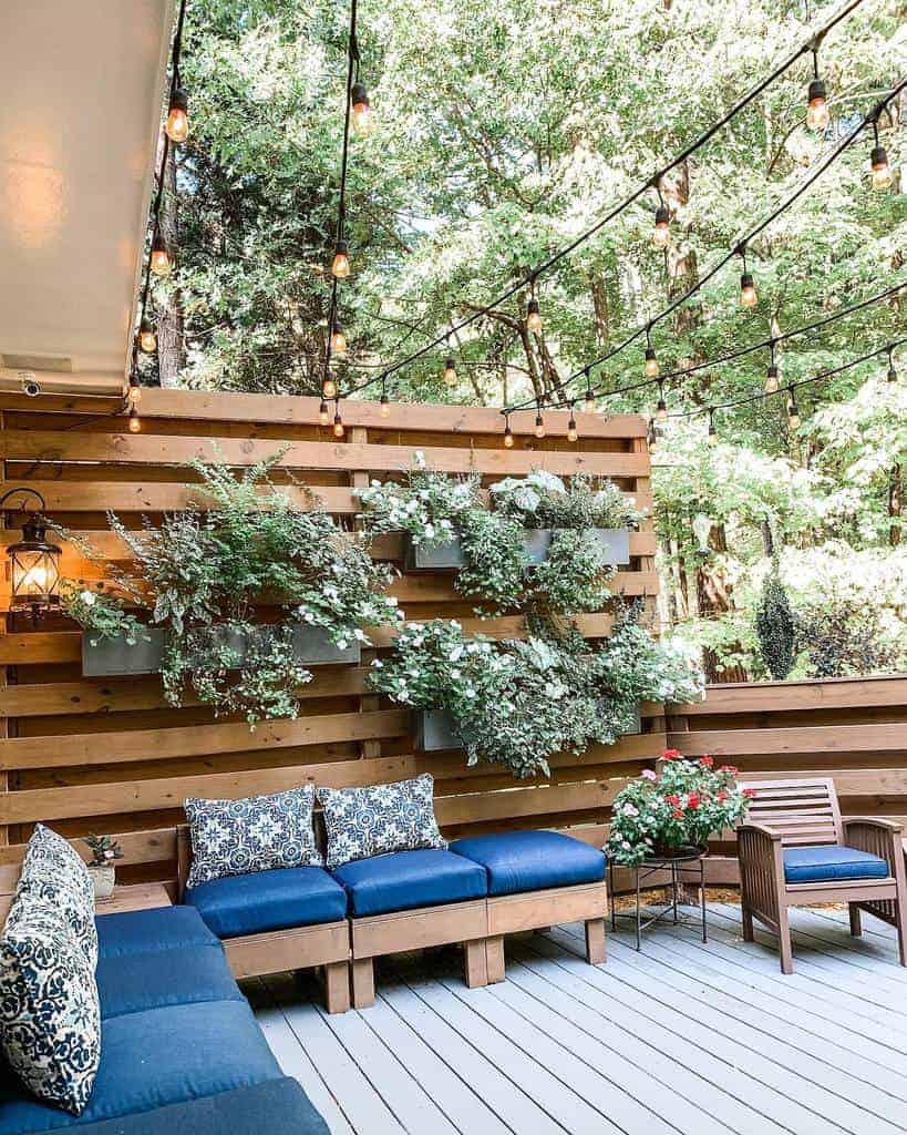 Cozy backyard patio with a wooden slat privacy fence, hanging greenery, blue cushioned seating, and string lights for a warm and inviting ambiance