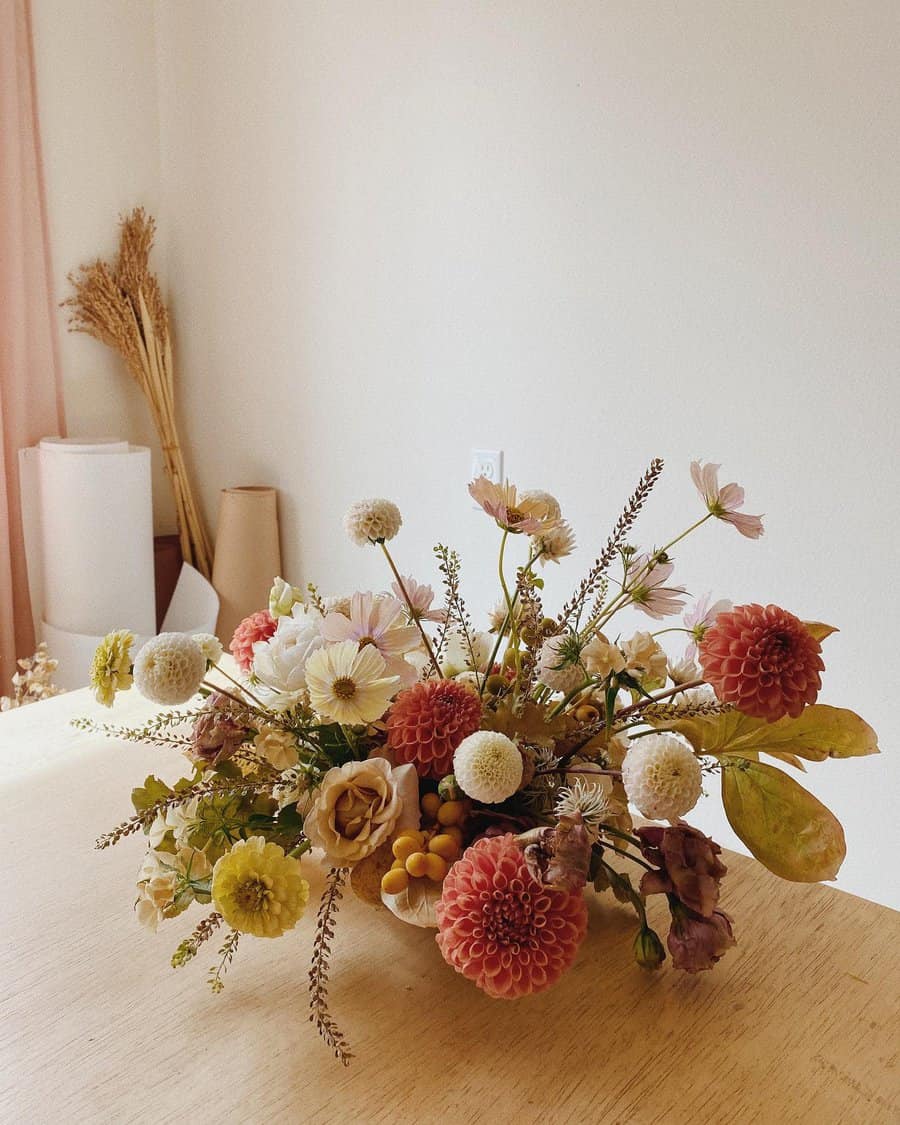 A floral arrangement with pink and white flowers and greenery sits on a wooden table in a minimalist room background