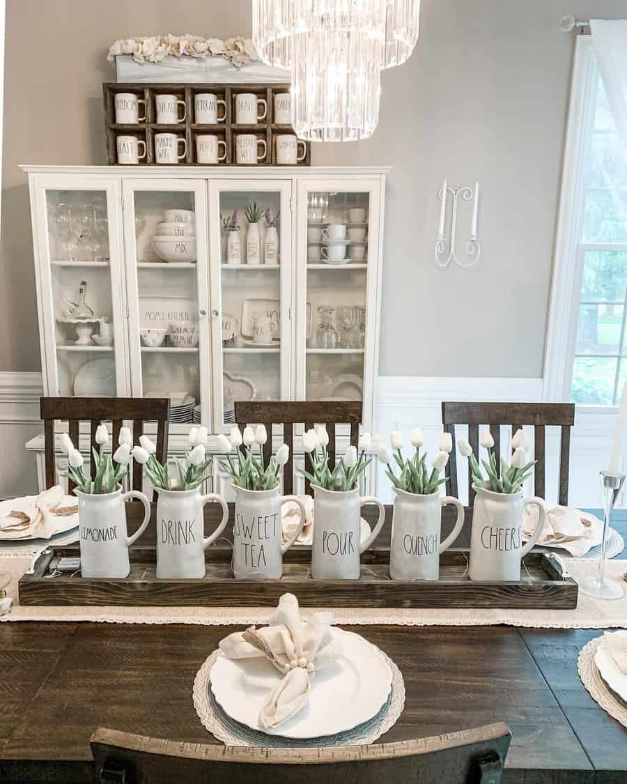 Dining room with a wooden table set for a meal, ceramic mugs with tulips are displayed as a centerpiece, and a cabinet in the background