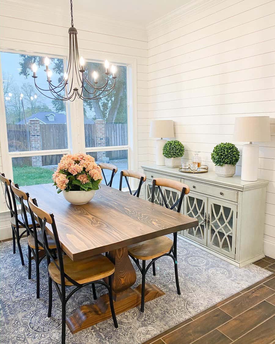 Bright farmhouse dining room with a rustic wood table, a floral centerpiece, black metal chairs, and a soft neutral-toned sideboard