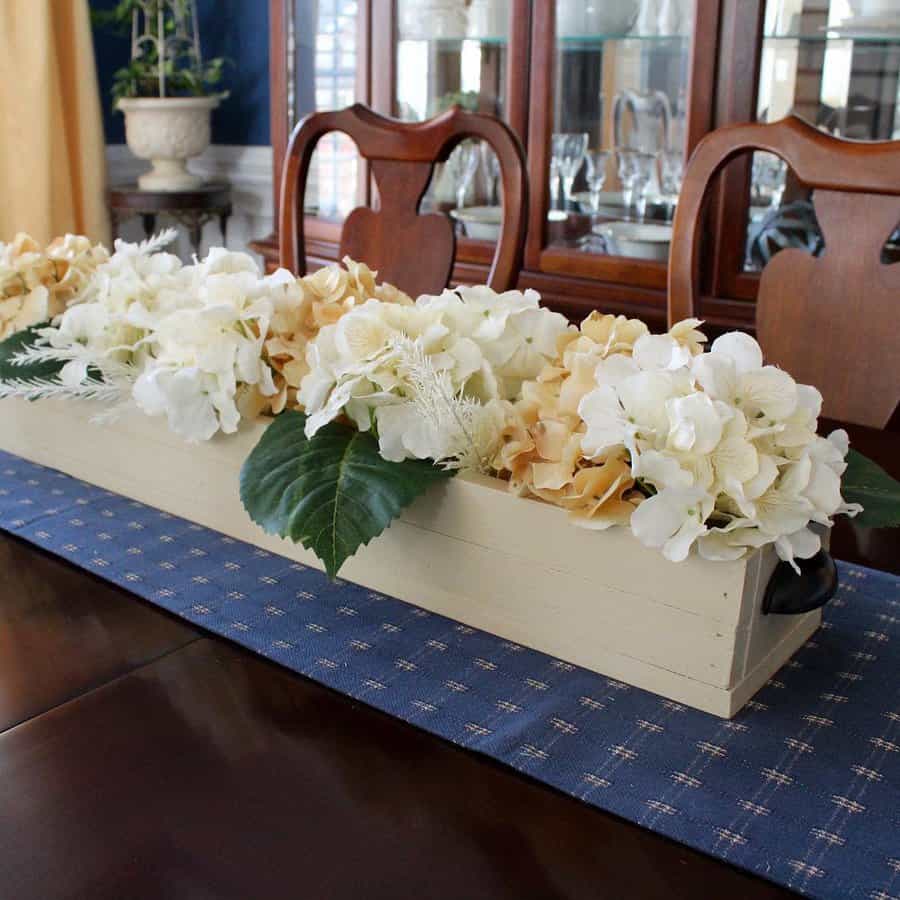 Rectangular wooden box with white and cream hydrangeas on a blue patterned table runner, set on a polished dining table