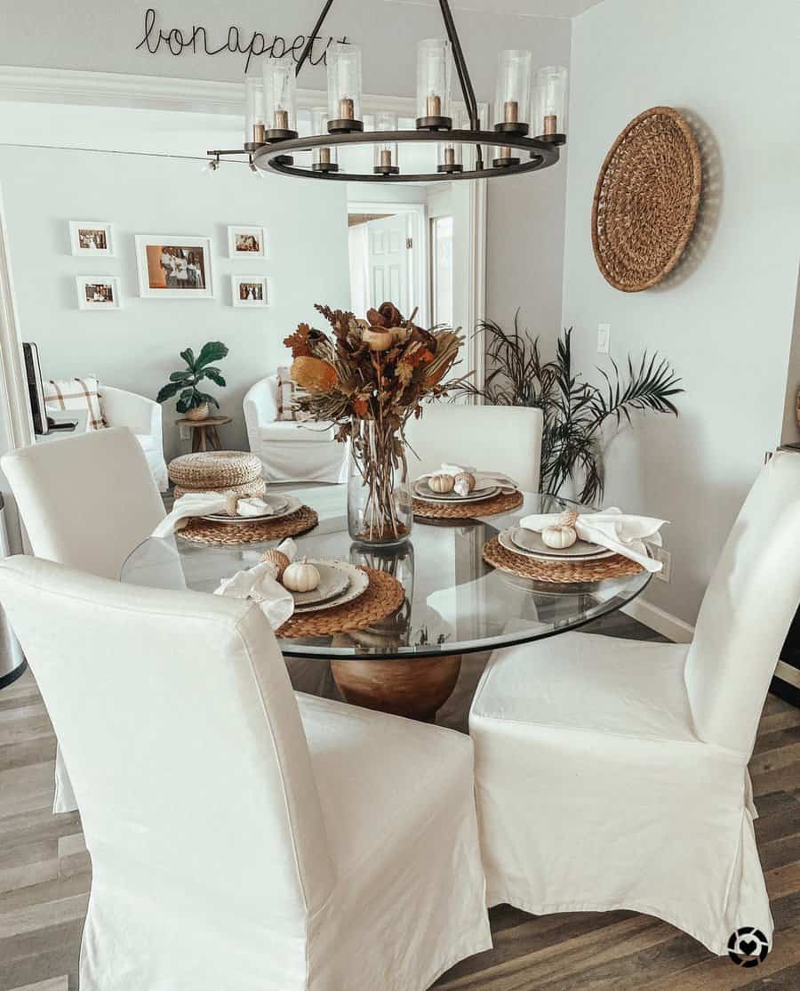 Cozy dining room with a glass table, white slipcovered chairs, woven placemats, and a fall-inspired floral centerpiece in a glass vase