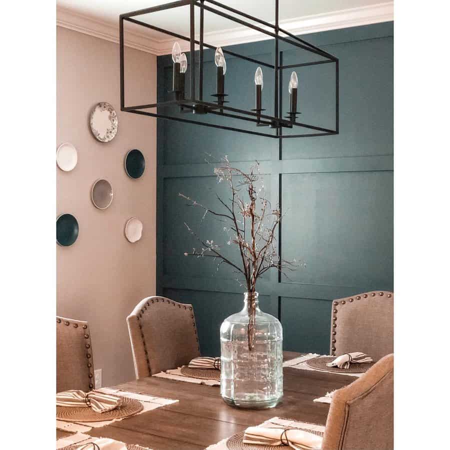 Dining room with a wooden table, beige chairs, a chandelier, decorative plates on a teal wall, and a large glass vase with branches