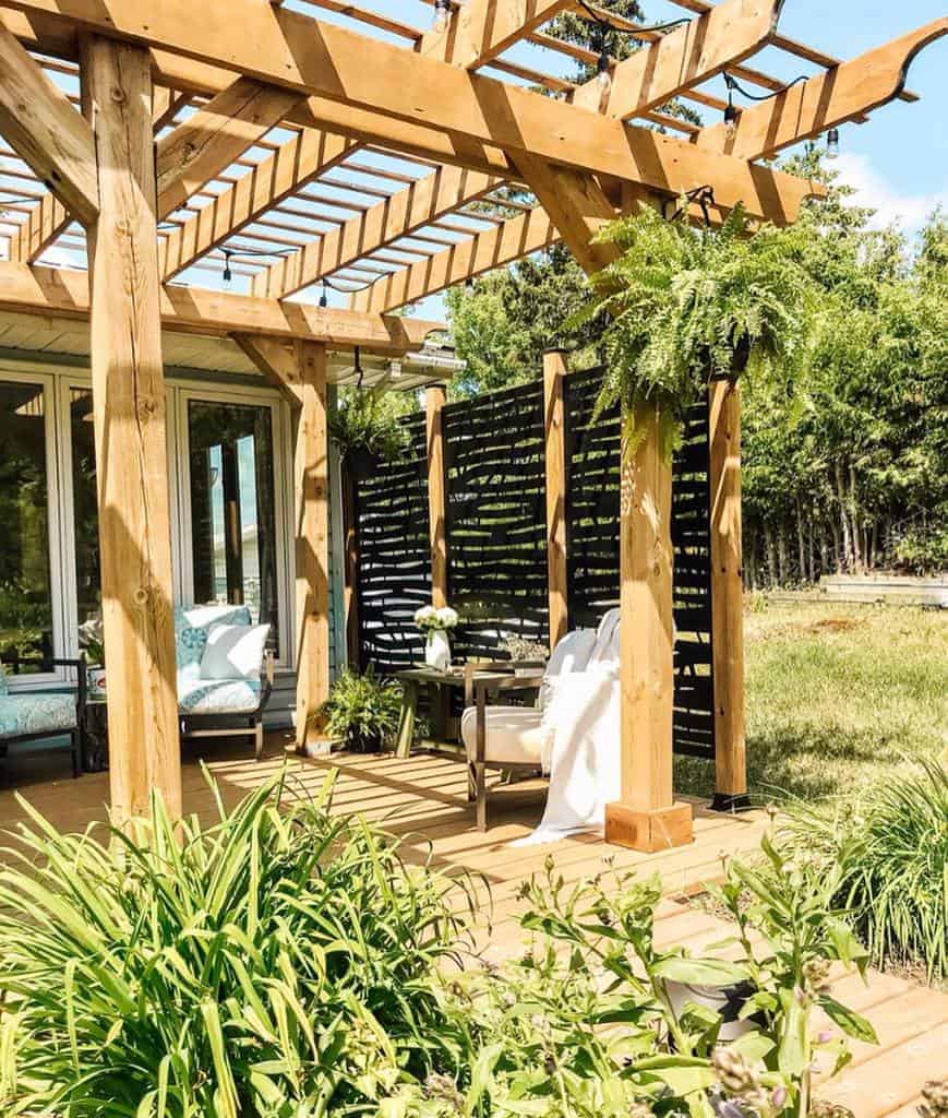 Beautiful outdoor pergola with a black slatted privacy screen, cozy seating, hanging ferns, and lush greenery, creating a relaxing backyard retreat