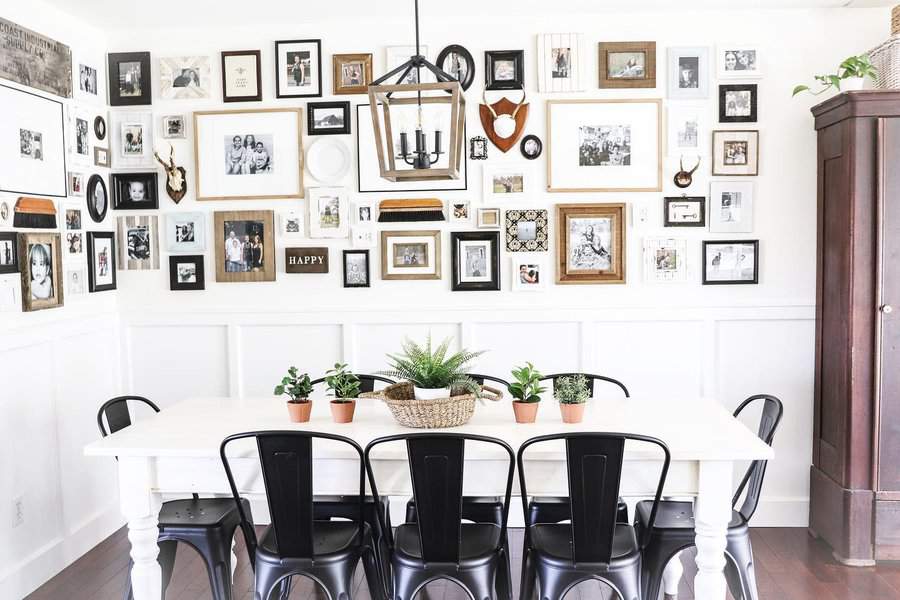 Bright farmhouse dining room with a white table, black metal chairs, a gallery wall of framed photos, and potted greenery centerpiece