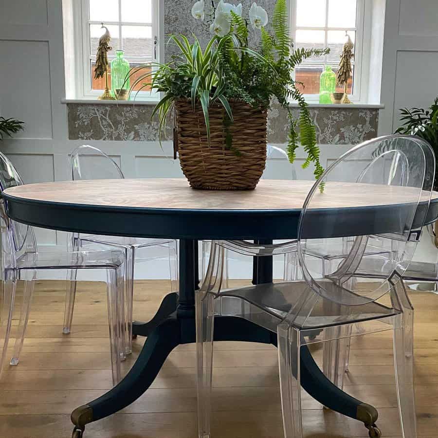 Round table with a wicker plant basket on top, surrounded by clear chairs, in a room with two windows and wooden flooring